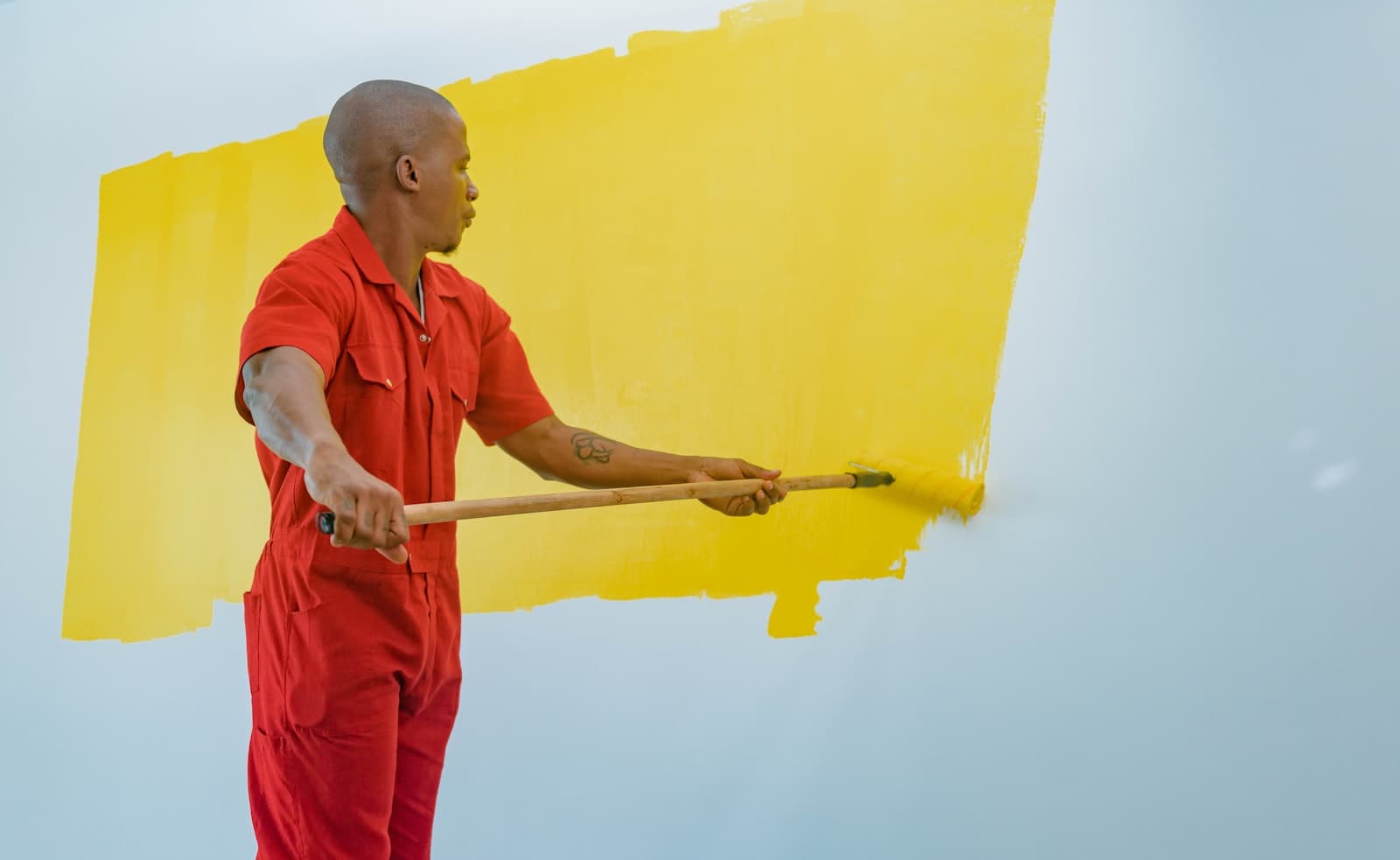A decorator in red overalls rolling fresh yellow paint onto a white wall
