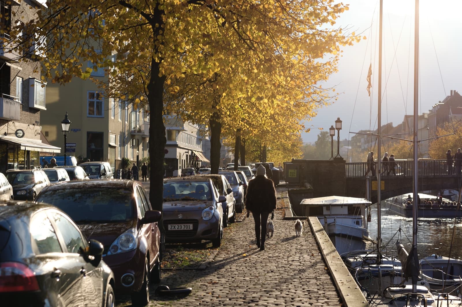 Golden autumn light on the cobbled canal-side quay of Christianshavn, Copenhagen, with moored boats and a person walking a dog beside the water.