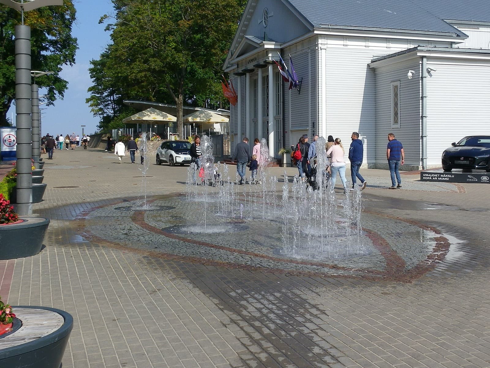 The plaza and fountain in front of the Dzintari Concert Hall on a sunny day, with café terraces and Latvian flags, Dzintari, Jūrmala, Latvia.