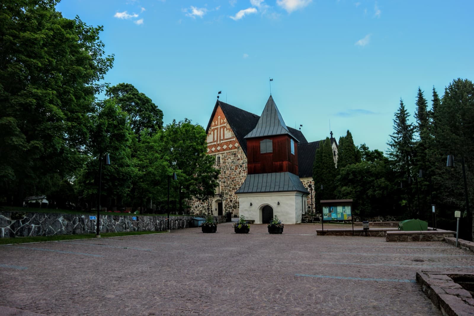The medieval greystone Espoo Cathedral with its red brick gable and wooden bell tower, seen across the cobbled square in Espoon keskus on a summer evening.