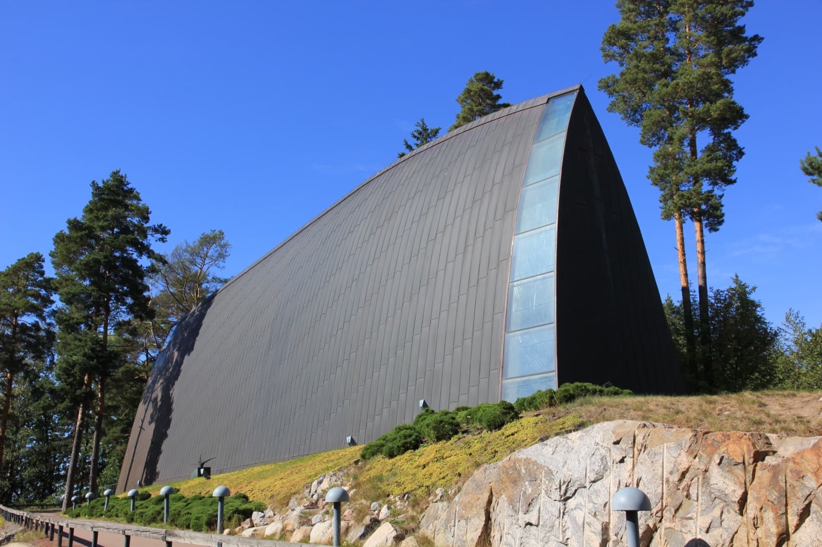 The curved wooden hull of St Henry's Ecumenical Art Chapel on its rocky pine ridge in Hirvensalo, Turku, under a blue sky.