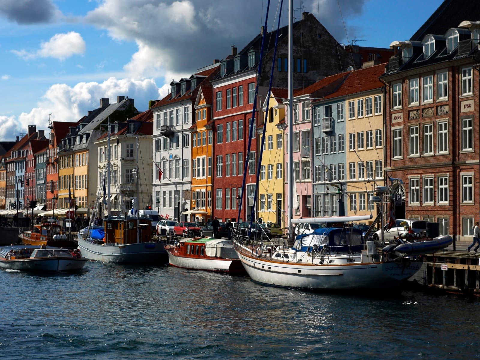 The colourful gabled townhouses and moored wooden sailboats of Nyhavn harbour in Indre By, Copenhagen, under a bright cloud-dappled sky.