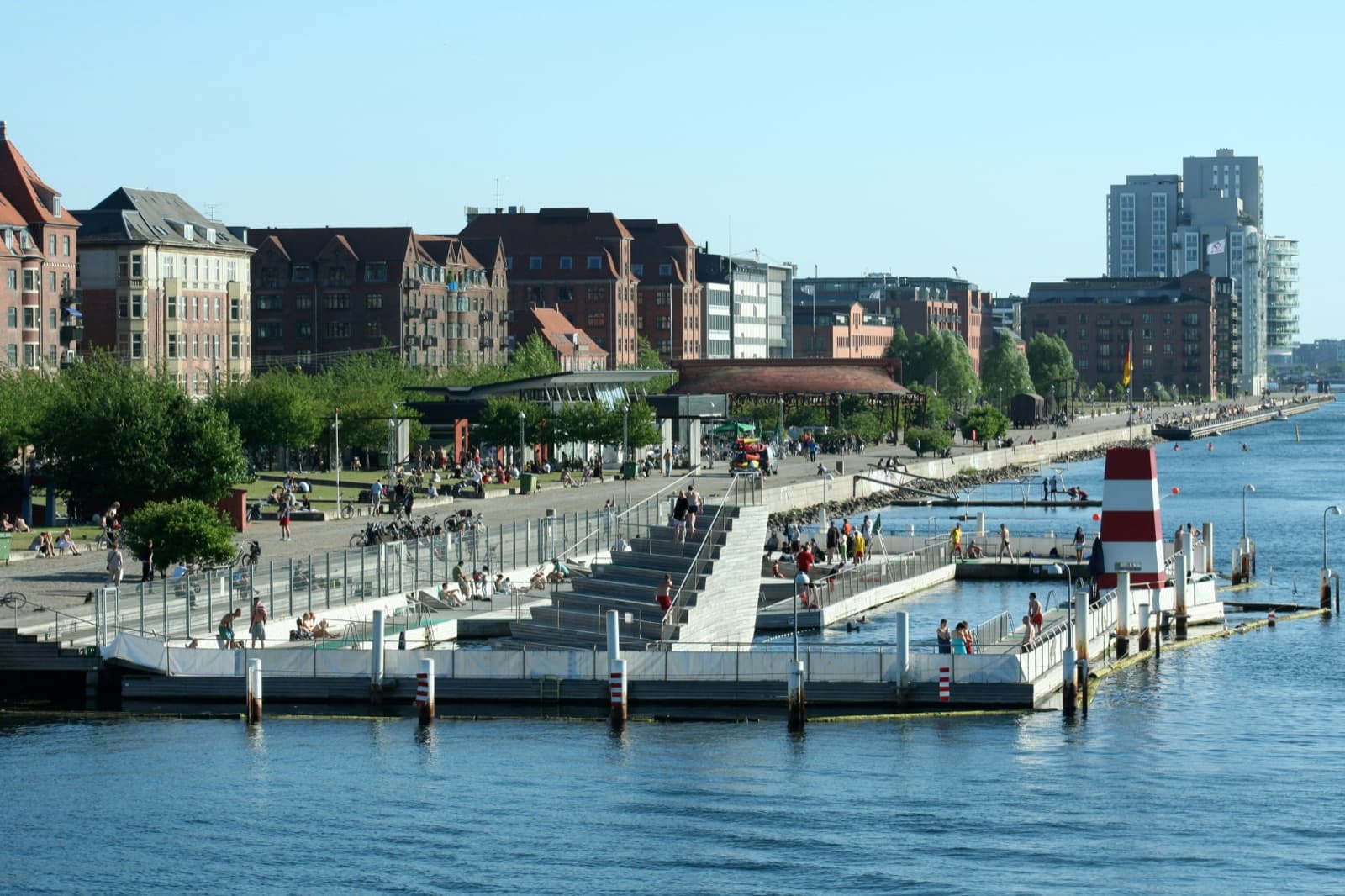 The Islands Brygge harbour bath and waterfront promenade in Copenhagen on a sunny day, with people relaxing along the quay and the harbour park in front of the district's buildings.