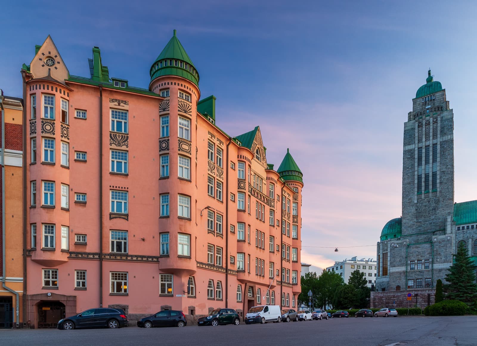 The pink Jugendstil Ihantola apartment building with green turrets on Linjat in Kallio, Helsinki, with the granite tower of Kallio Church beside it at sunset.