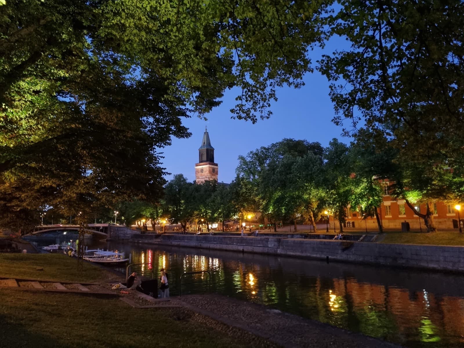 The Aura river at dusk in central Turku, lamps reflected in the water and the tower of Turku Cathedral rising behind the riverside trees.