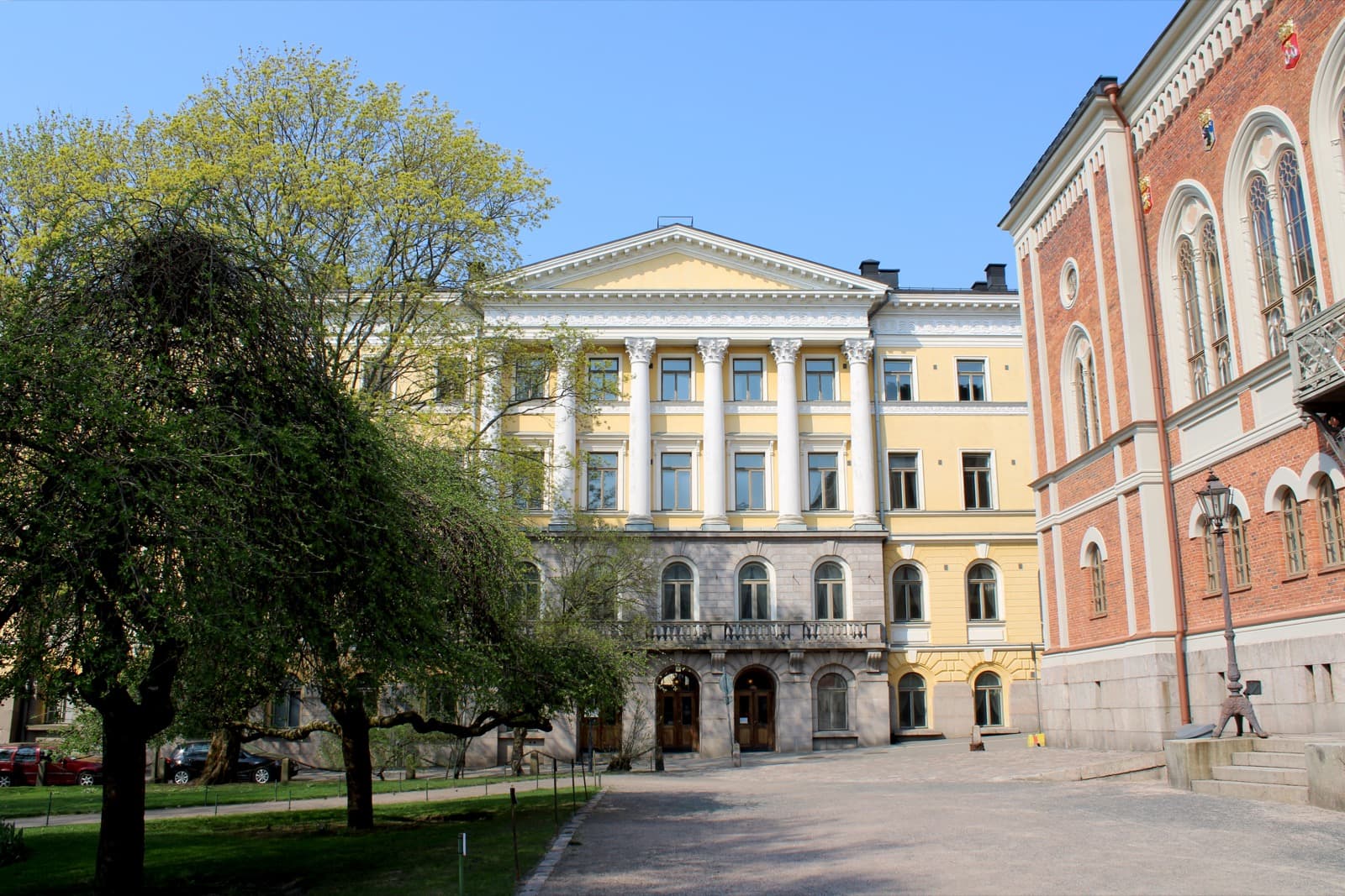 The yellow neoclassical Government Palace seen from the park beside the red brick House of Nobility on Ritarikatu in Kruununhaka, Helsinki, under a clear spring sky.