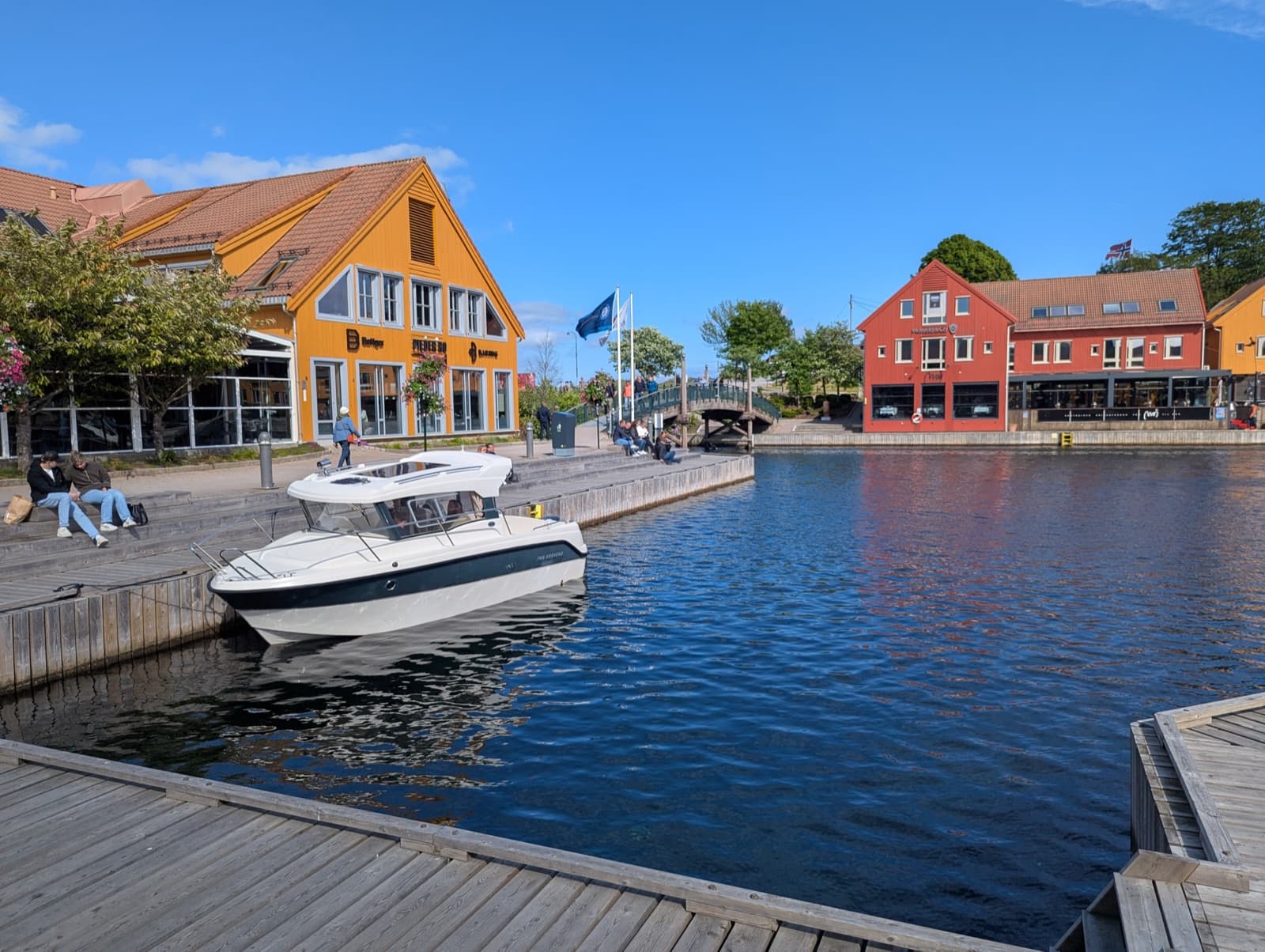 A boat moored on the canal at Fiskebrygga, with yellow and red timber quayside buildings under a blue sky, Kvadraturen, Kristiansand, Norway.