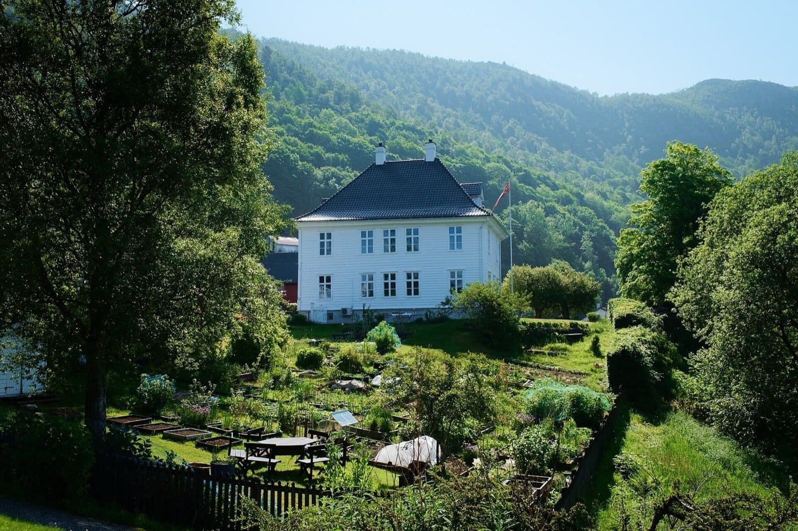 The white timber Landås Hovedgård manor in Bergen, with allotment beds in front and the wooded slope of Landåsfjellet behind.