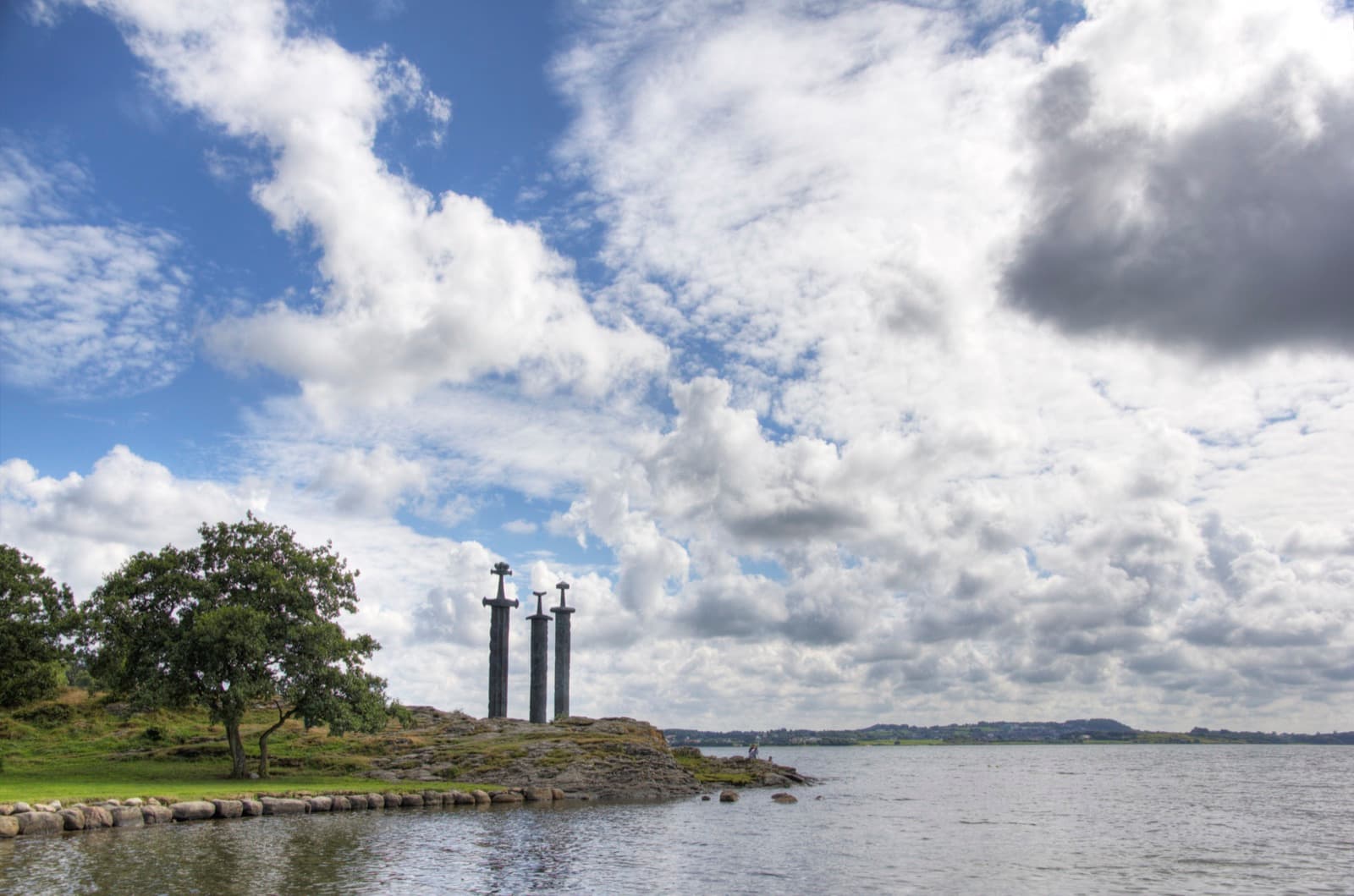 The three Sverd i fjell swords standing on the rocky shore of Hafrsfjord at Møllebukta under a big cloudy sky, Madla, Stavanger, Norway.