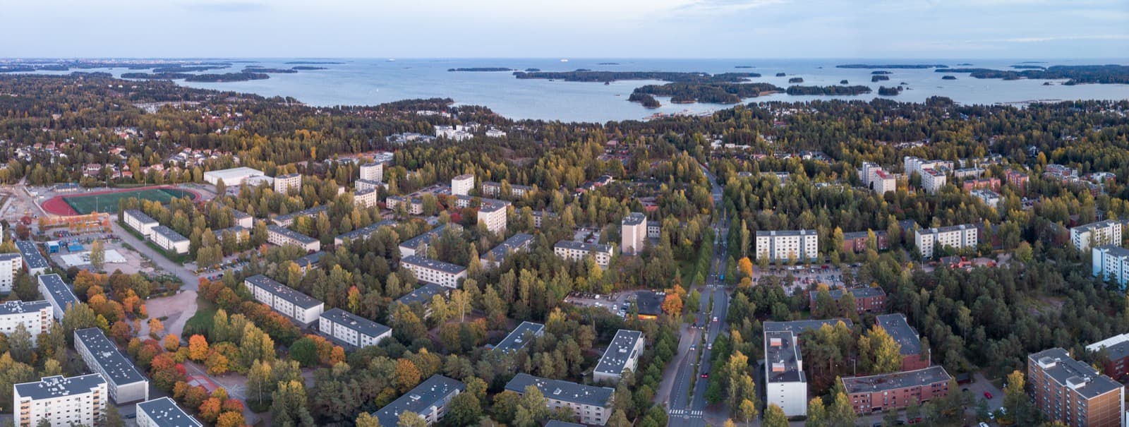 An aerial panorama of Matinkylä in Espoo at dusk, low apartment blocks set among autumn trees running down to the Gulf of Finland and its islands.