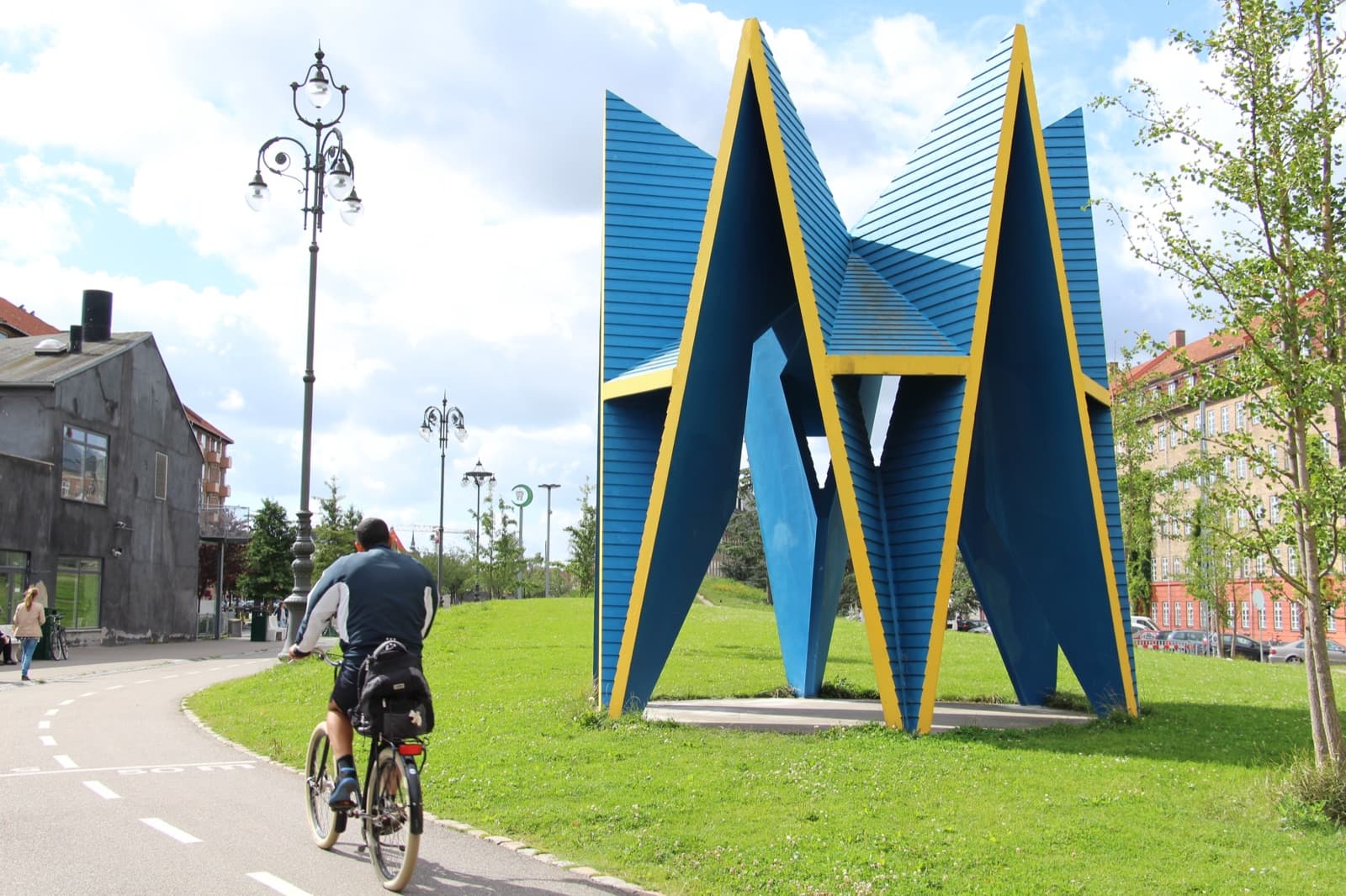 The blue and yellow sculptural pavilion in the Superkilen park in Nørrebro, Copenhagen, with a cyclist passing on the bike path and an ornate lamppost nearby.