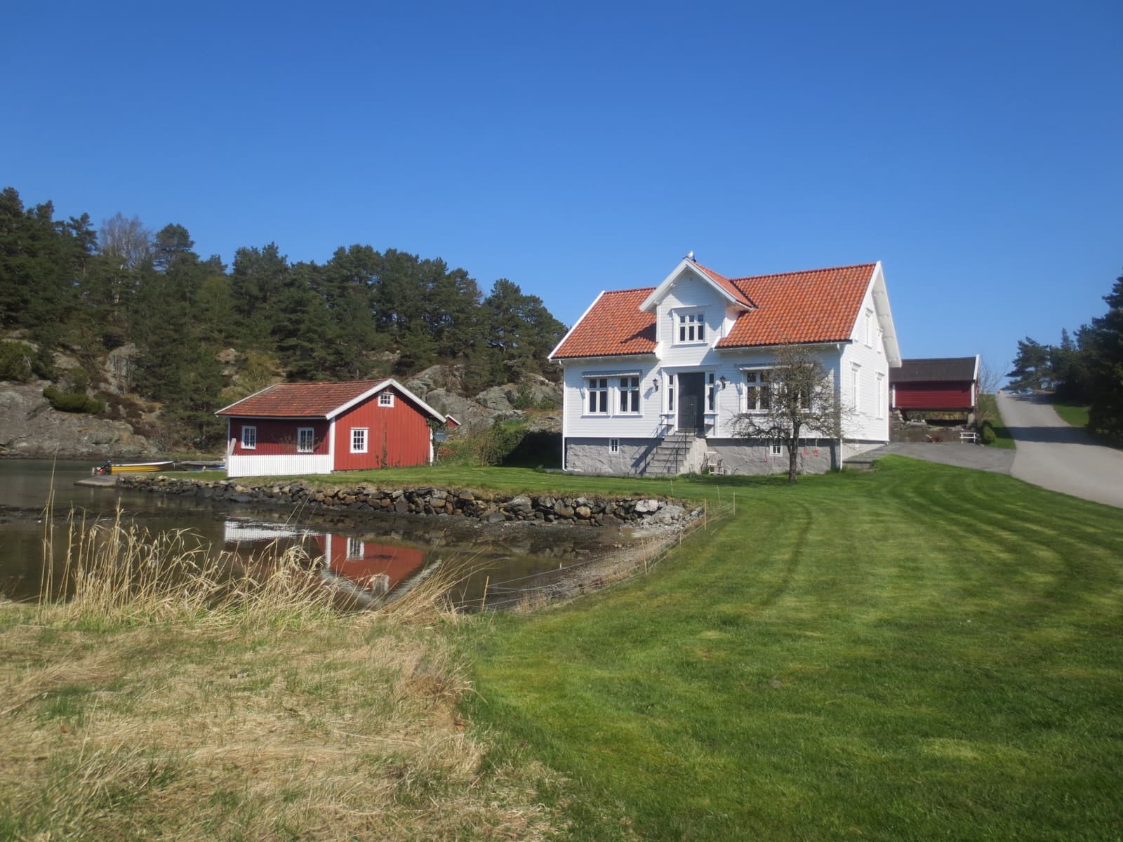 A white timber shore farmhouse and red boathouse beside a sheltered inlet at Kvarenes, Randesund, Kristiansand, Norway.