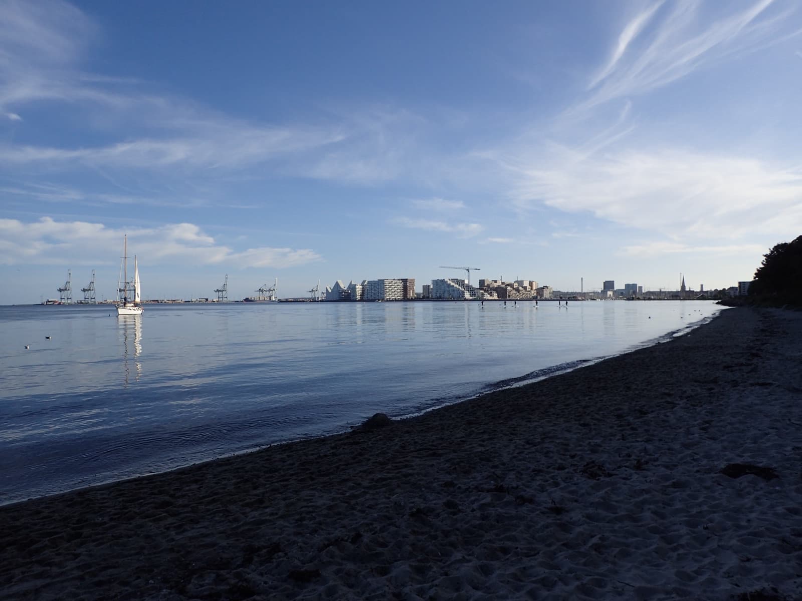 The beach at Den Permanente in Risskov, Aarhus, looking across the bay to the city skyline with a sailboat.