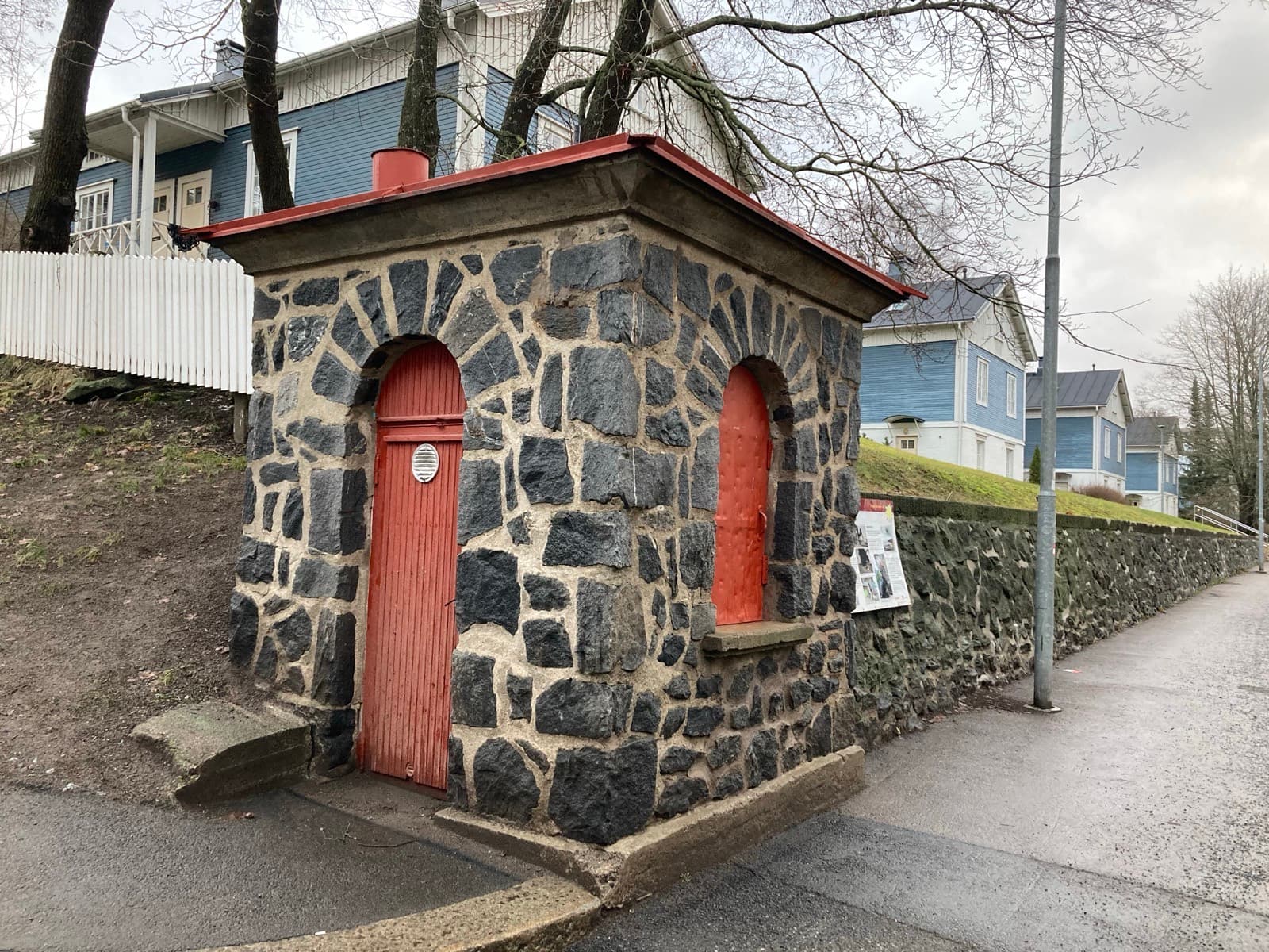 The little stone kiosk on Annikinkatu in Tammela, Tampere, with the blue wooden houses of the Annikki block behind it.