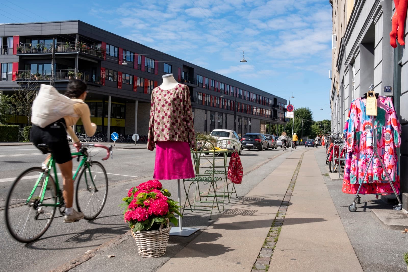 Street life on Valby Langgade in Copenhagen, with a cyclist passing a shop's outdoor clothing and flower display under a blue sky.