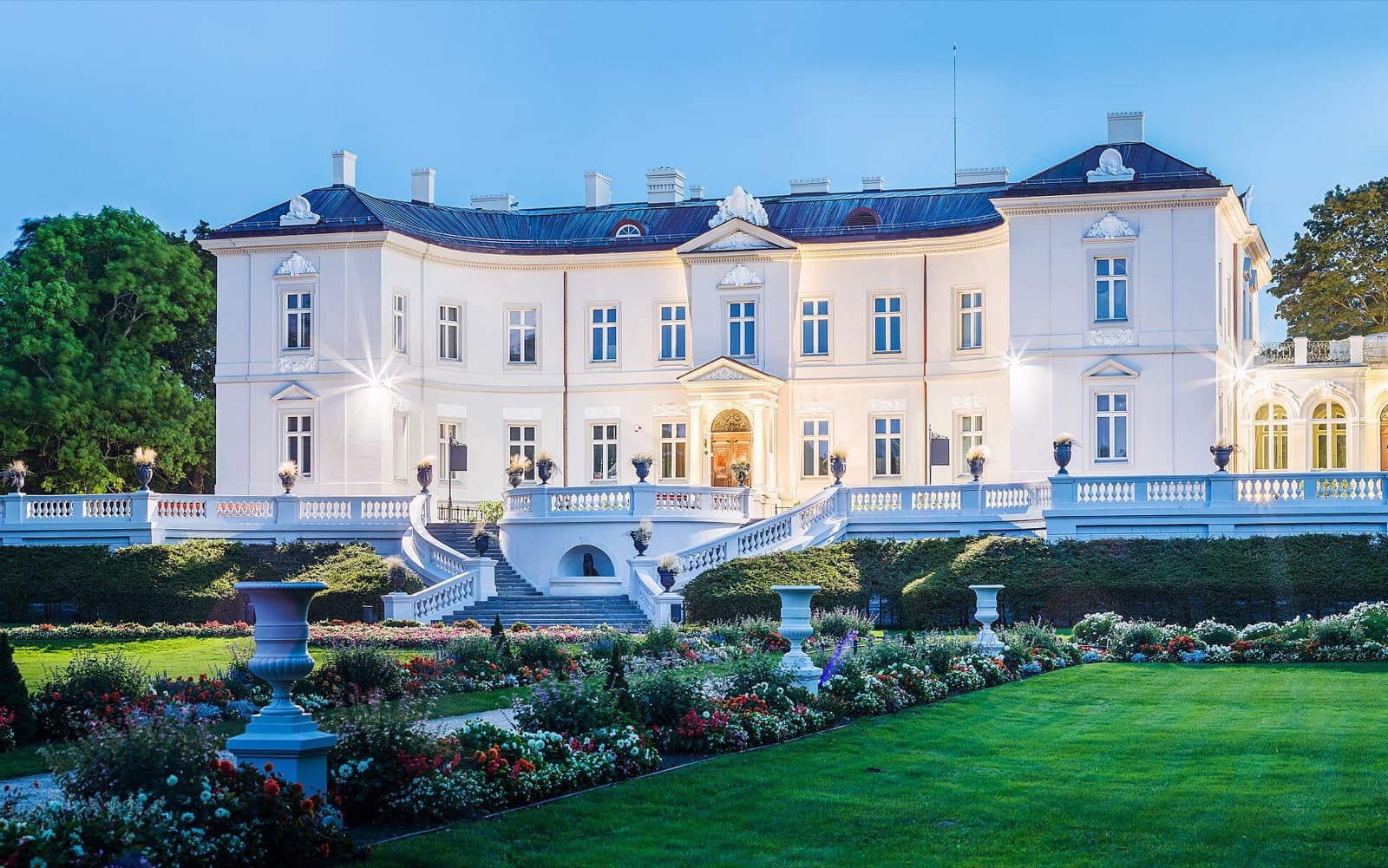 The white neoclassical Tiškevičiai Palace, now the Amber Museum, lit at dusk above its formal flower garden in the Palanga Botanical Park