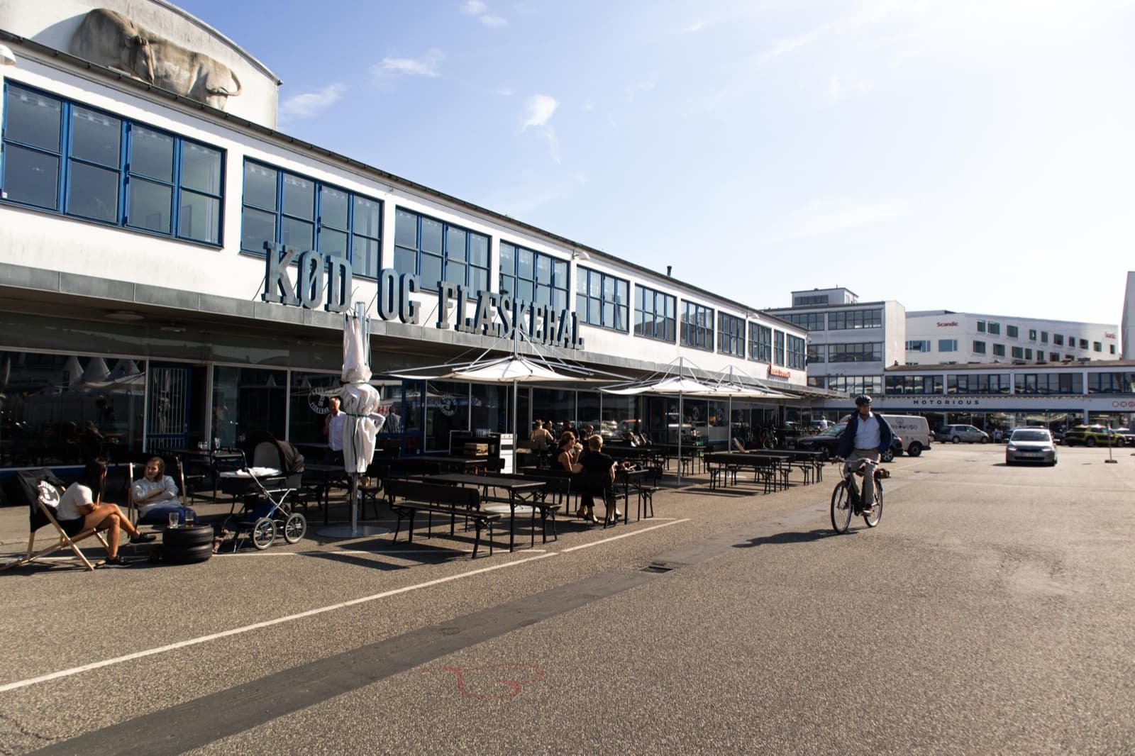 The white functionalist halls of the Meatpacking District (Kødbyen) in Vesterbro, Copenhagen, with outdoor café seating and a cyclist in warm evening light.