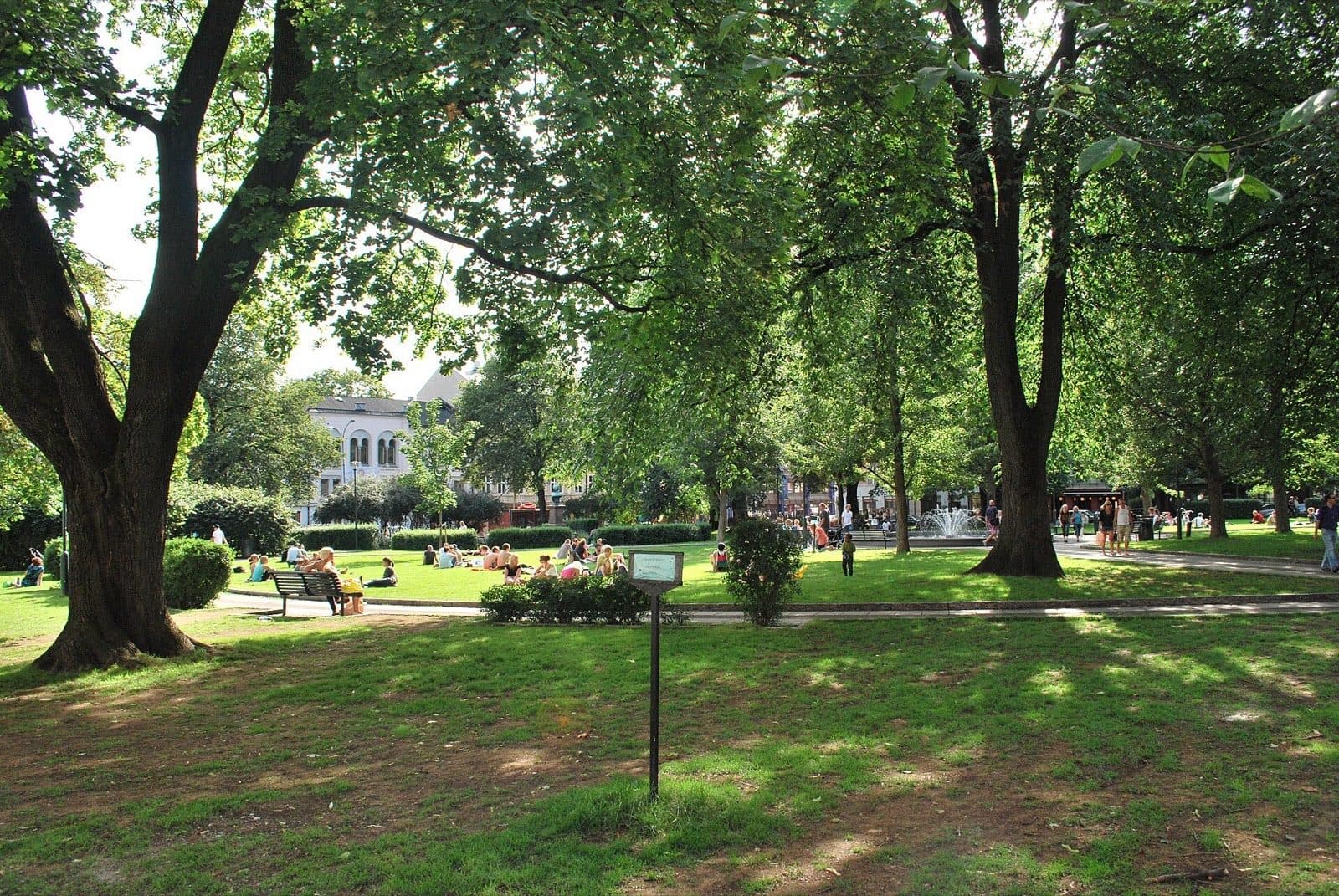 Olaf Ryes plass in Grünerløkka, Oslo, on a summer day, with people sitting on the grass under mature trees and the fountain behind.