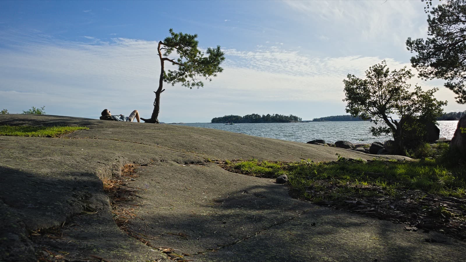 Smooth shoreline rocks on Lauttasaari island in Helsinki, with a leaning pine, someone sunbathing and the sea and wooded islets beyond.