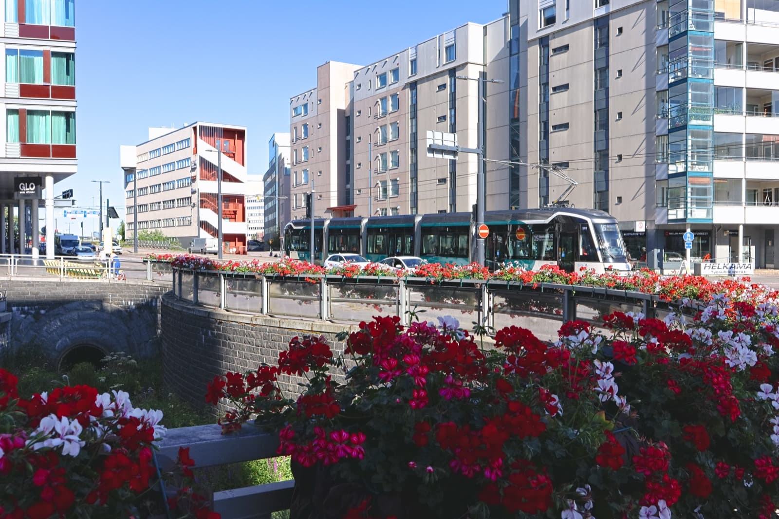 A light rail tram passing modern apartment blocks in the centre of Leppävaara, Espoo, with beds of red and white geraniums in the foreground on a clear summer day.