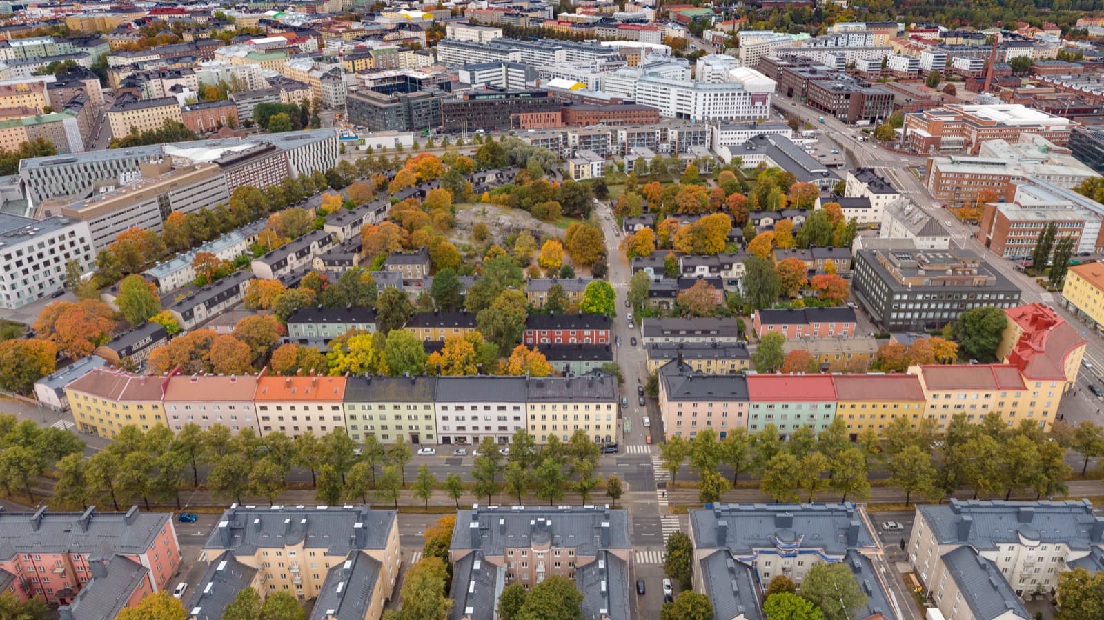 An aerial view over Puu-Vallila in Helsinki, the low painted wooden houses and pastel apartment terraces set among autumn trees.